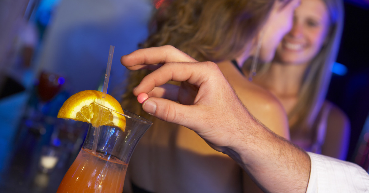 image depicting a hand dropping drugs into an alcoholic drink. Woman are seen in the background