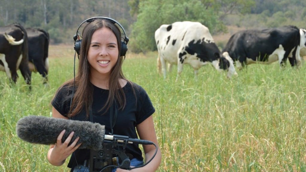 Alexandra Green at Mayfarm holding a microphone. Black and white cows in a grassy field can be seen behind her. 